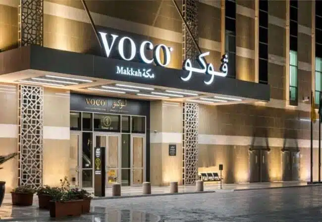 The entrance of Voco Makkah Hotel, featuring illuminated signage, large windows, and contemporary design elements.